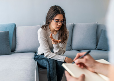 Young woman in a therapy session expressing concern while a therapist takes notes.