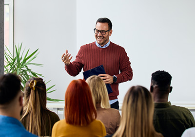 A man in a red sweater speaking to an audience in a bright room.