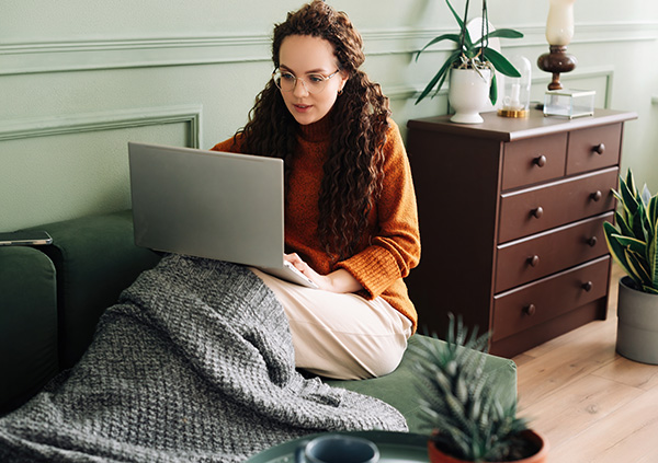 Woman in an orange sweater sitting on a couch with a laptop, wrapped in a blanket.