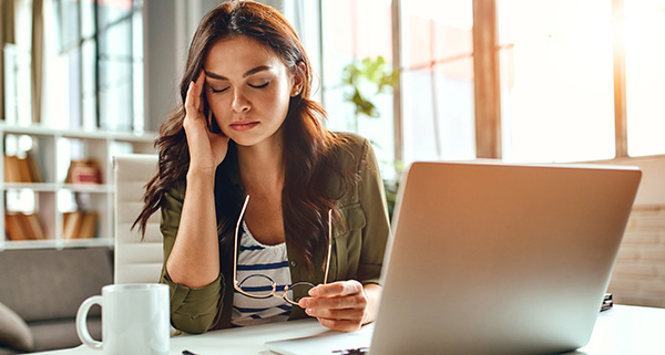 Woman sitting at a desk with a laptop, appearing stressed while holding her glasses.