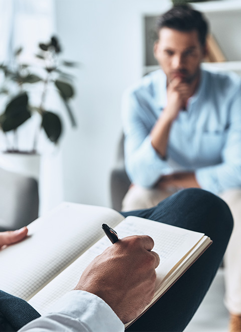 Client seated in a therapy session while therapist takes notes in a notebook.