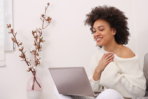 Smiling woman in cozy sweater using a laptop for online therapy session.