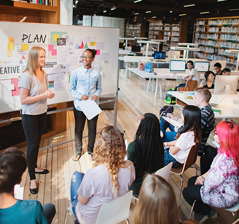 Two women presenting in a spacious, modern classroom with students attentively listening.